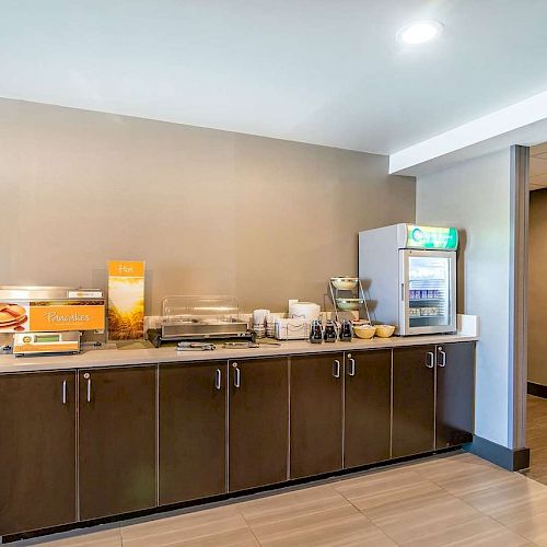 The image shows a hotel breakfast bar featuring a juice dispenser, coffee machines, bread, and a cereal dispenser, with cupboards below.