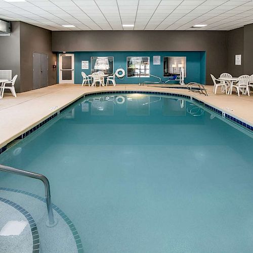 An indoor swimming pool with surrounding deck chairs and tables, situated in a well-lit room with a tiled ceiling and ventilation ducts.