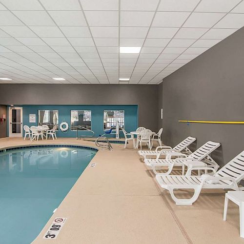 An indoor pool area with lounge chairs, white tables, and safety equipment on the walls. The pool is clean and the deck looks well-maintained.