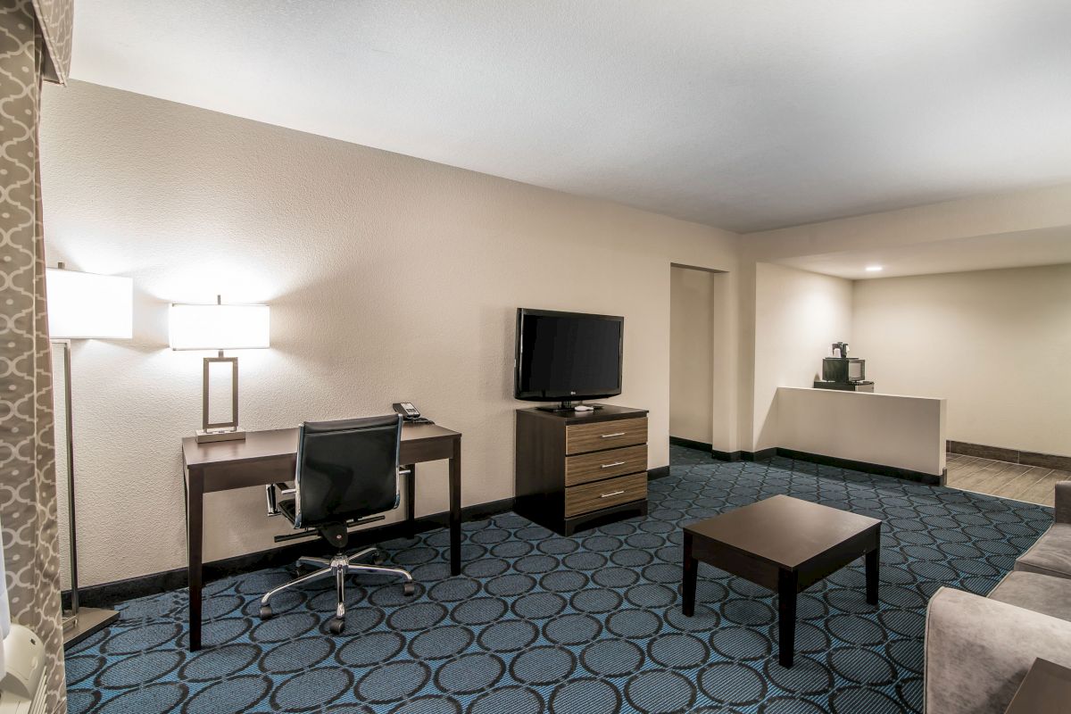 A hotel room features a desk with a lamp and chair, a TV on a dresser, and a coffee station on a counter, all on a blue-patterned carpet.