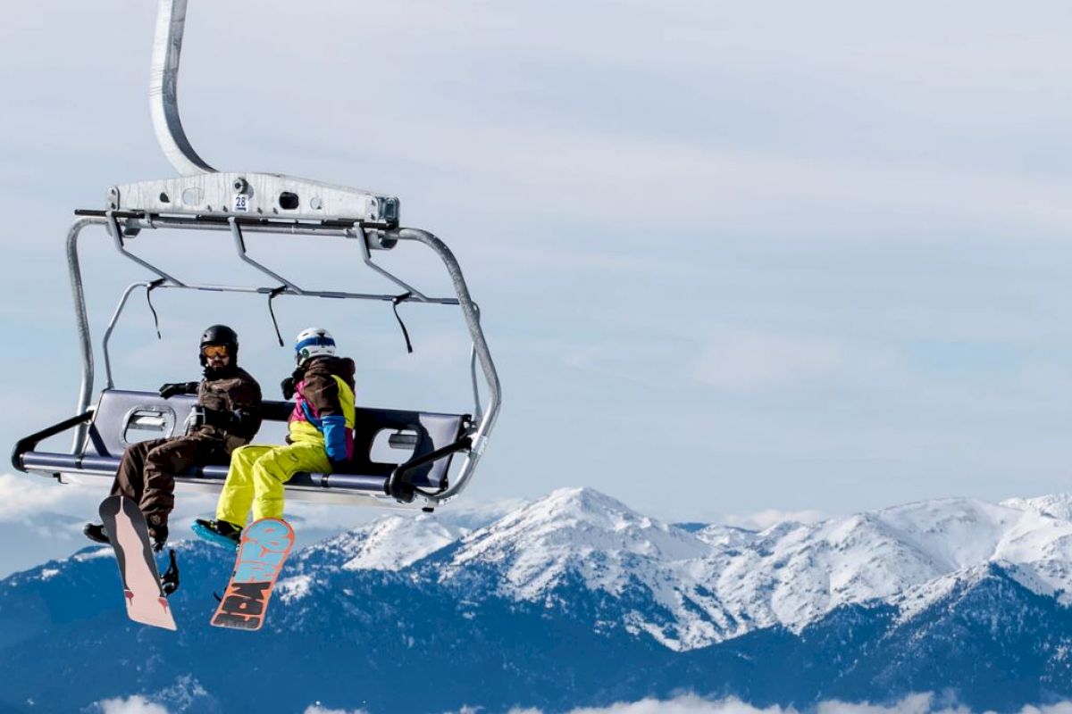 Two people are on a ski lift with snow-covered mountains in the background during a winter day.