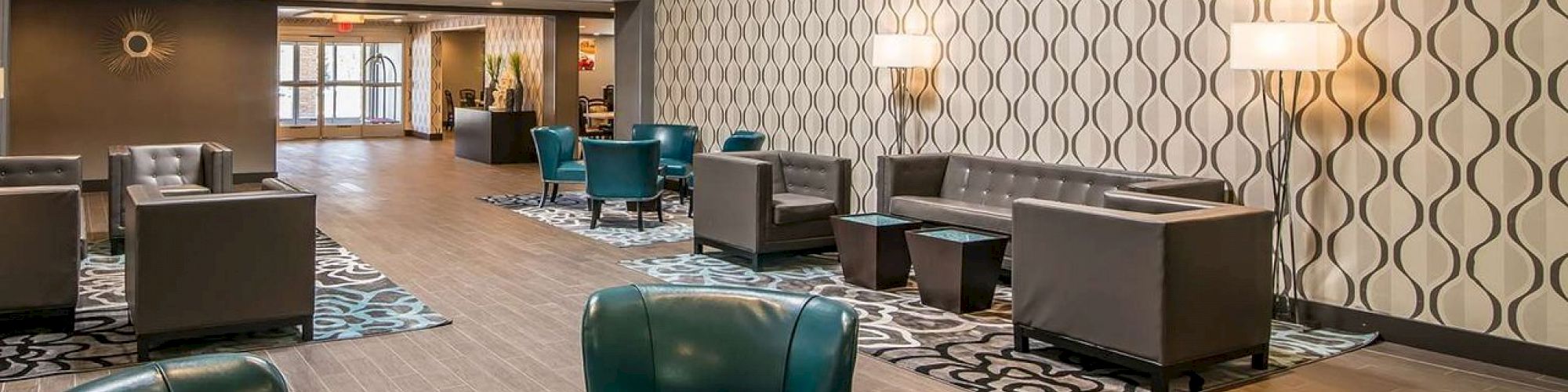 A modern hotel lobby with teal chairs, patterned carpet, and textured walls. The space has ample seating, a high ceiling, and ambient lighting.